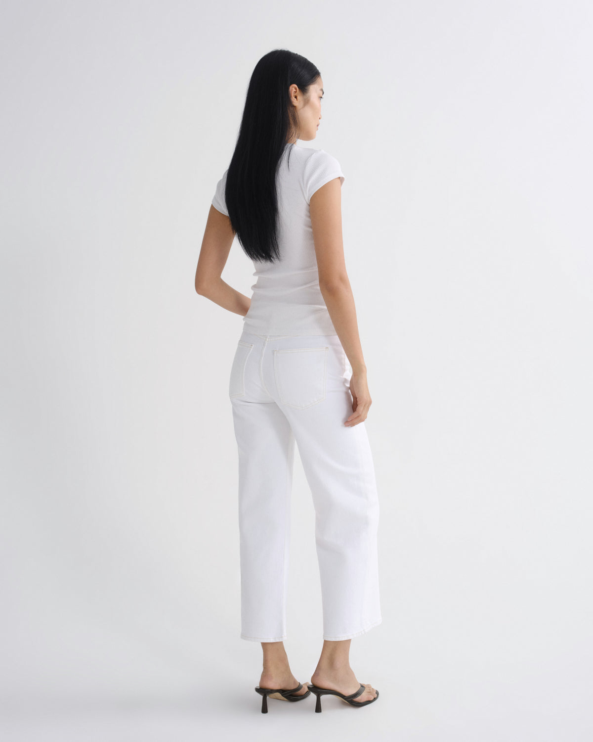 A woman with long black hair is shown from the back wearing a white short-sleeve top and white cropped pants, paired with black heeled sandals, against a plain white studio background.