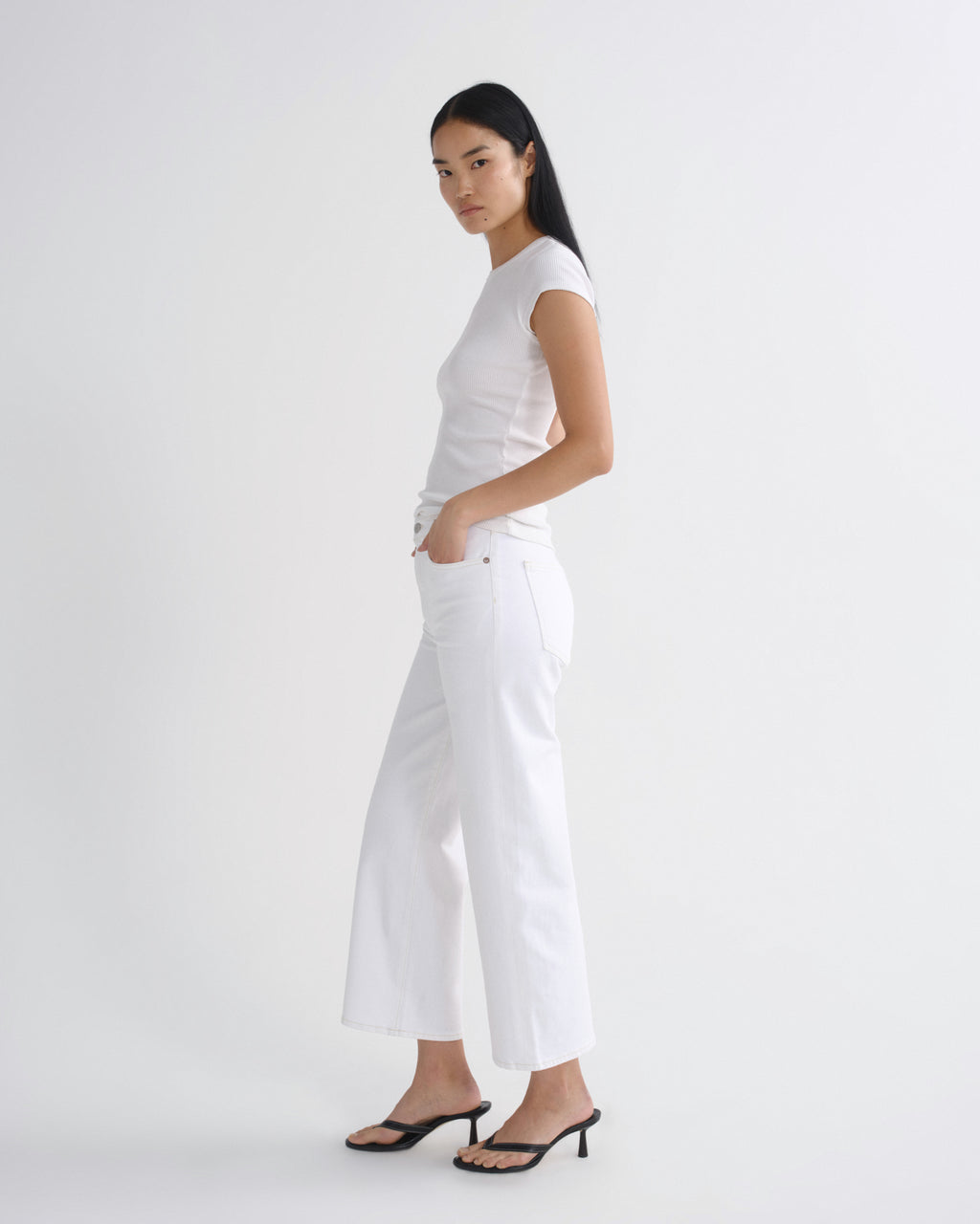 A woman modeling a white short-sleeve top and white wide-leg cropped pants, paired with black heeled sandals, against a plain white studio background.
