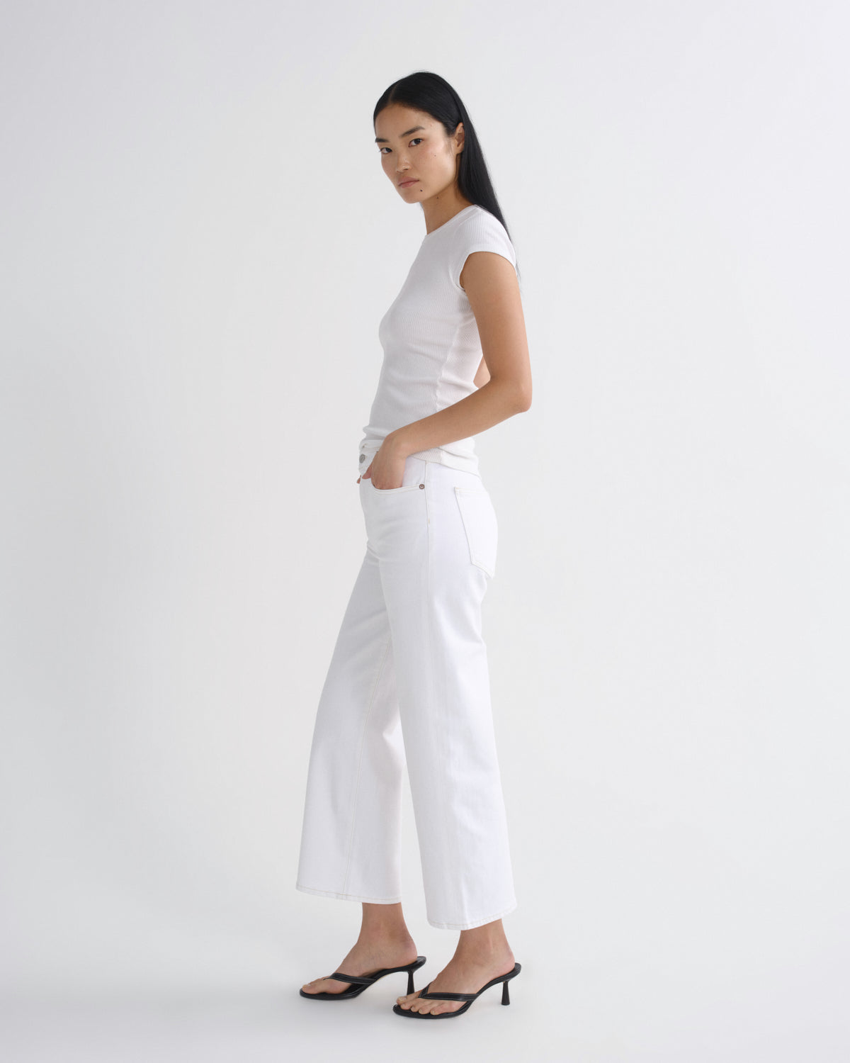 A woman modeling a white short-sleeve top and white wide-leg cropped pants, paired with black heeled sandals, against a plain white studio background.