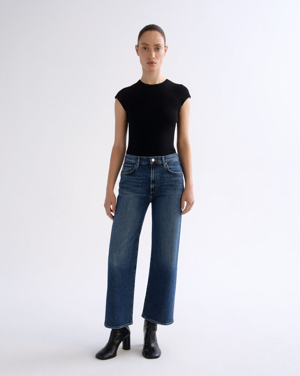 A model stands against a plain white studio background wearing a fitted black short-sleeve top, blue wide-leg jeans, and black ankle boots.