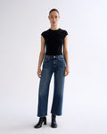 A model stands against a plain white studio background wearing a fitted black short-sleeve top, blue wide-leg jeans, and black ankle boots.