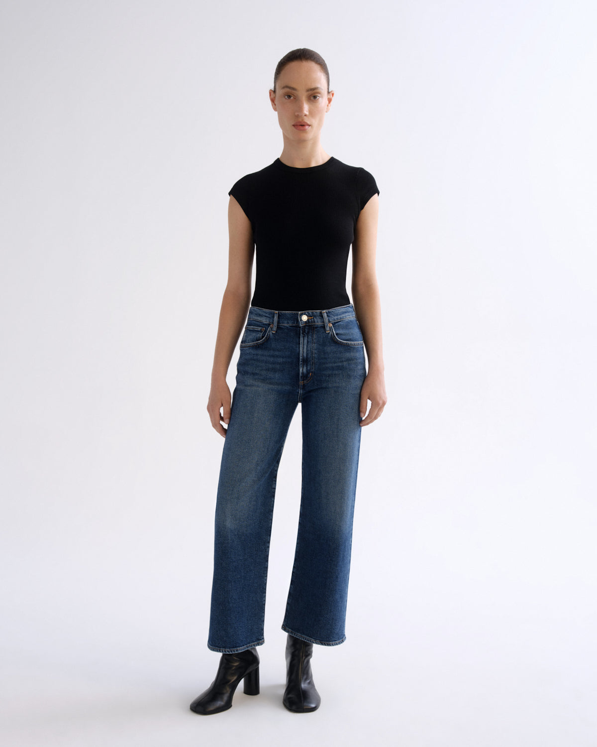 A model stands against a plain white studio background wearing a fitted black short-sleeve top, blue wide-leg jeans, and black ankle boots.