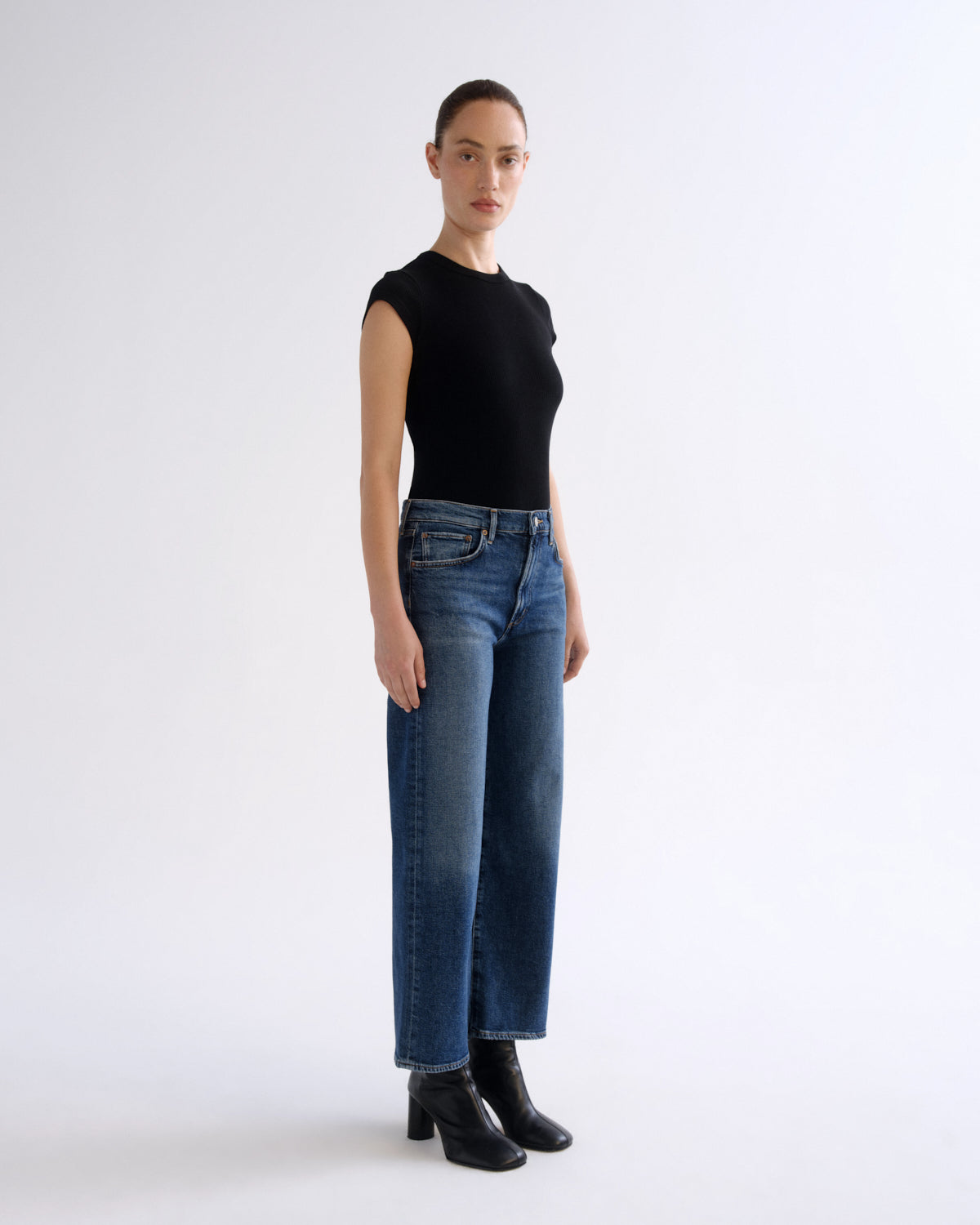 A model stands in profile wearing a black sleeveless top, blue wide-leg jeans, and black ankle boots against a plain light gray studio background.