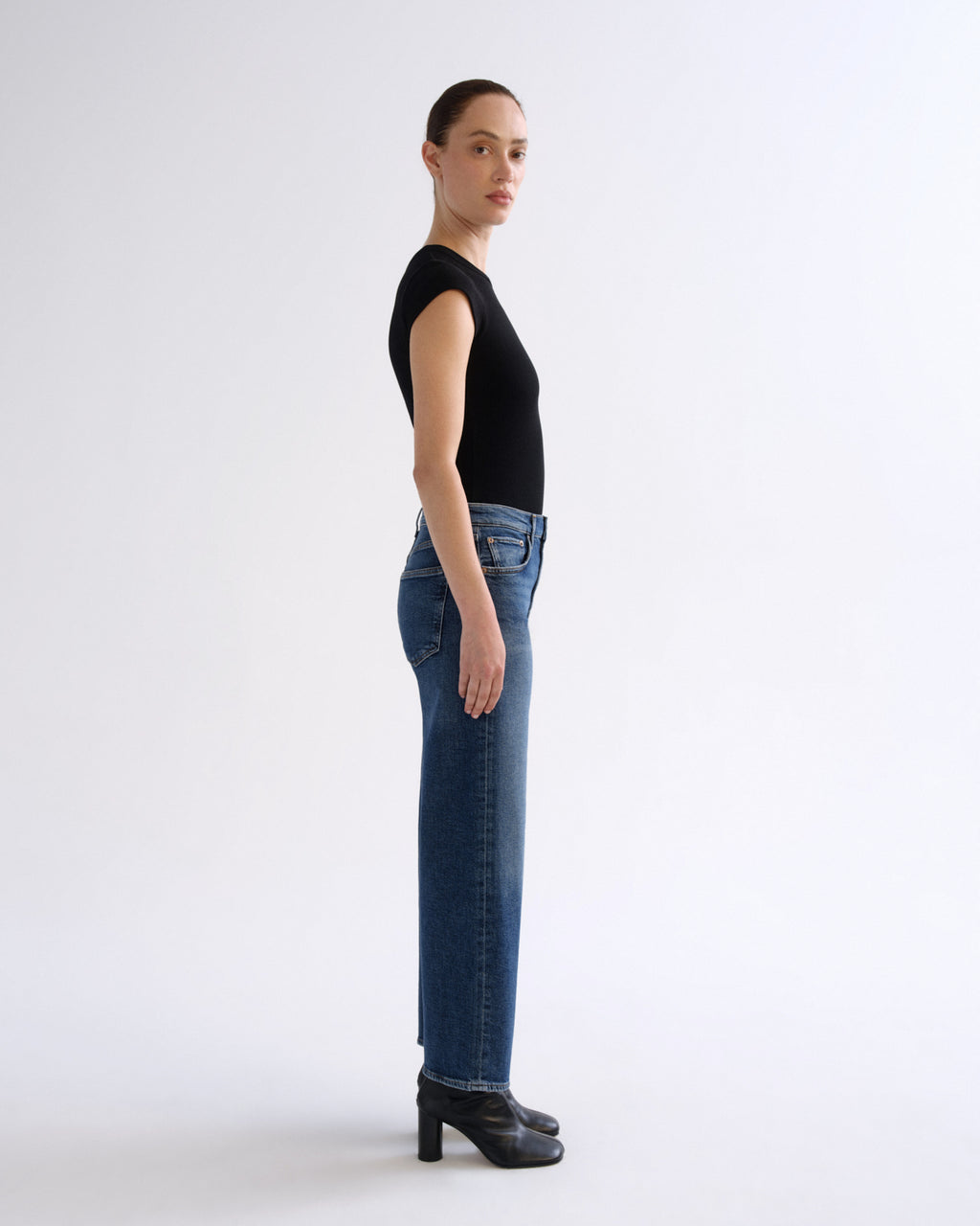 A model standing in profile wearing a black short-sleeve top, blue straight-leg jeans, and black heeled ankle boots against a plain light gray studio background.
