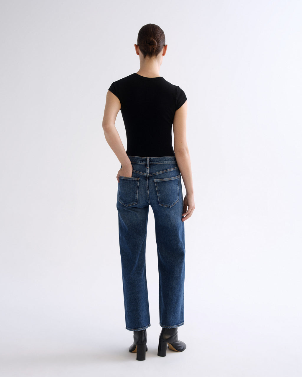 A model viewed from the back wearing a black short-sleeve top, blue straight-leg jeans, and black ankle boots against a plain white studio background.
