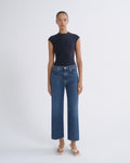 A model stands against a light gray studio background wearing a dark navy sleeveless mock-neck top and blue cropped wide-leg jeans with white thong sandals.