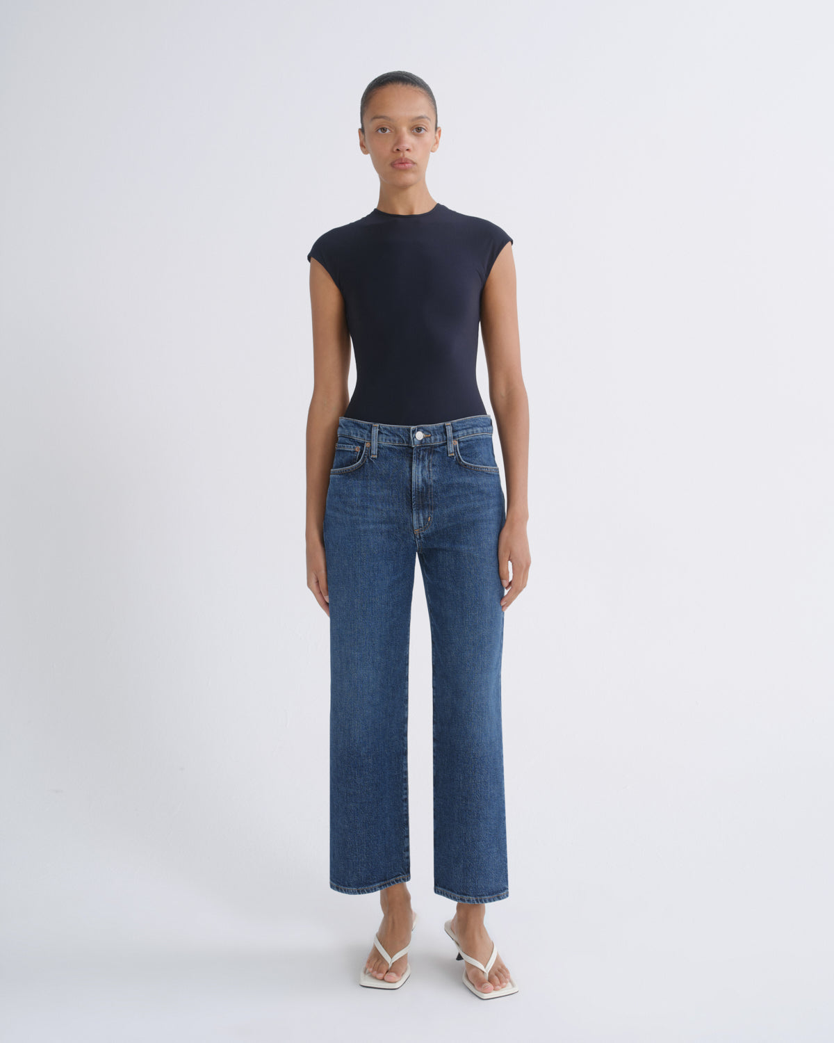 A model stands against a light gray studio background wearing a dark navy sleeveless mock-neck top and blue cropped wide-leg jeans with white thong sandals.