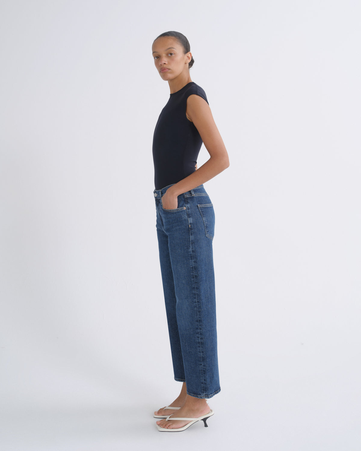 A model stands in profile wearing a black sleeveless top, blue wide-leg cropped jeans, and white heeled sandals against a plain light gray studio background.