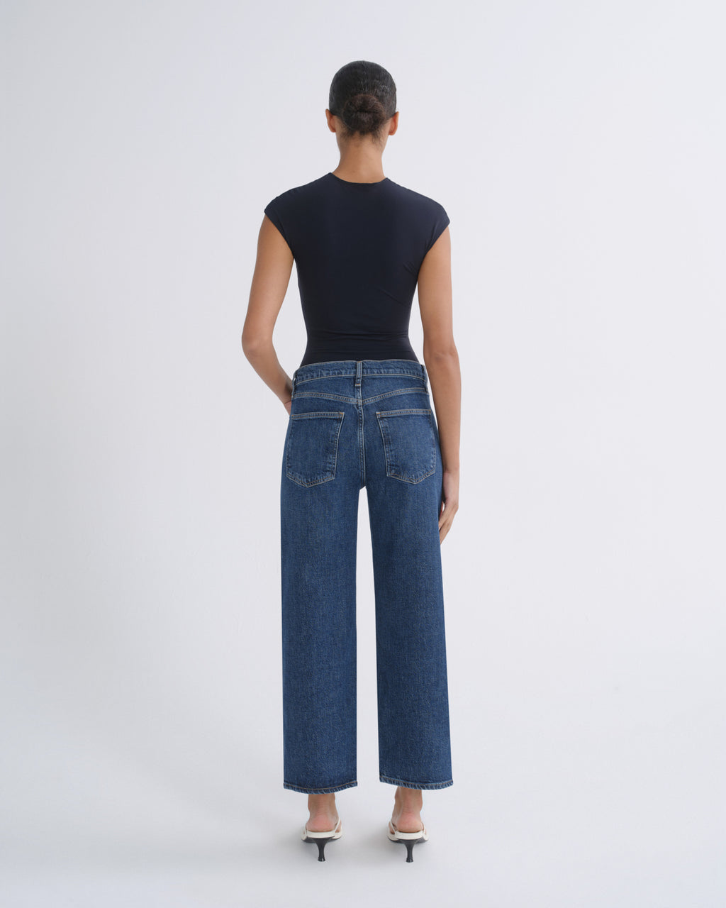 A model shown from the back wearing a dark navy sleeveless top, blue wide-leg cropped jeans, and white heeled sandals against a light gray studio background.