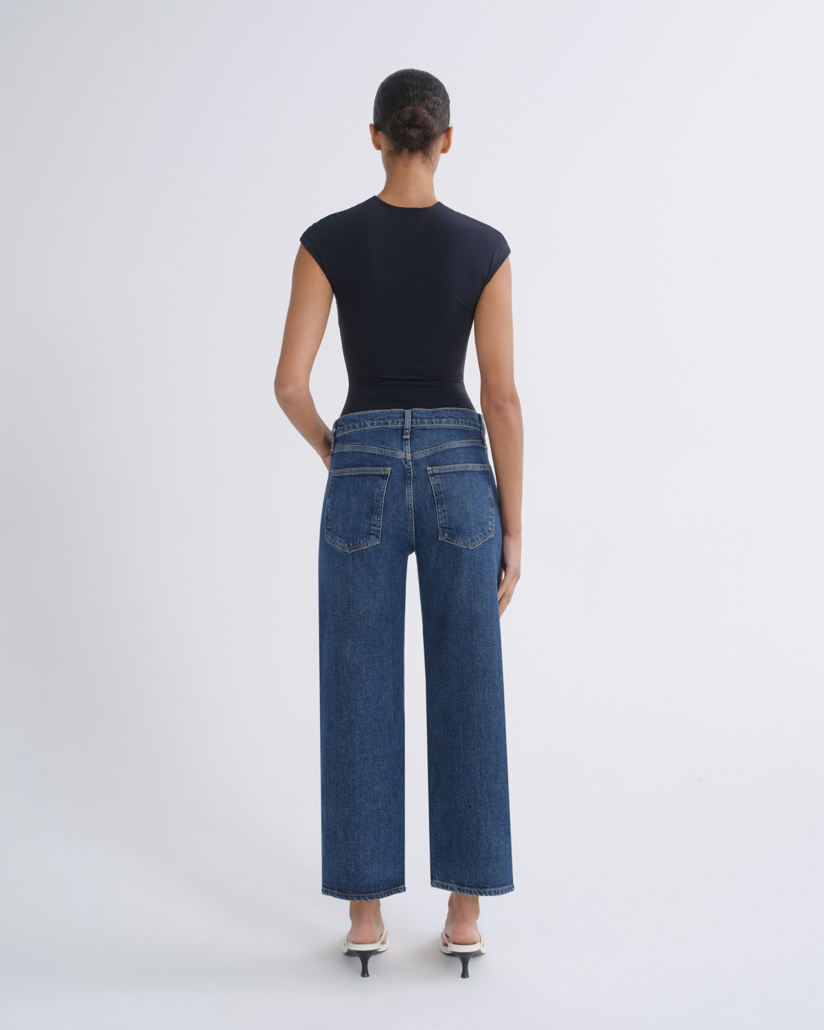 A model shown from the back wearing a dark navy sleeveless top, blue wide-leg cropped jeans, and white heeled sandals against a light gray studio background.