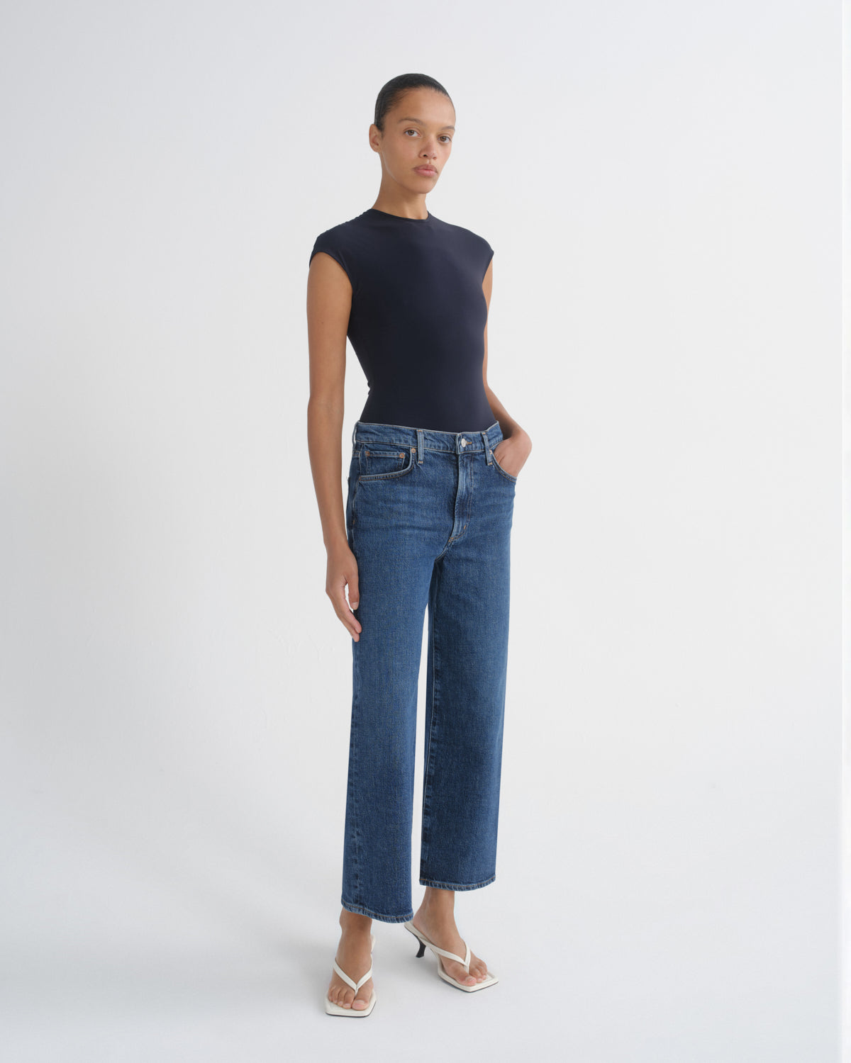 A model stands against a light gray studio background wearing a dark navy sleeveless top, blue straight-leg jeans, and white heeled sandals.