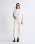 A model stands against a light gray studio background wearing a white short-sleeve t-shirt tucked into off-white wide-leg jeans, paired with dark brown ankle boots.