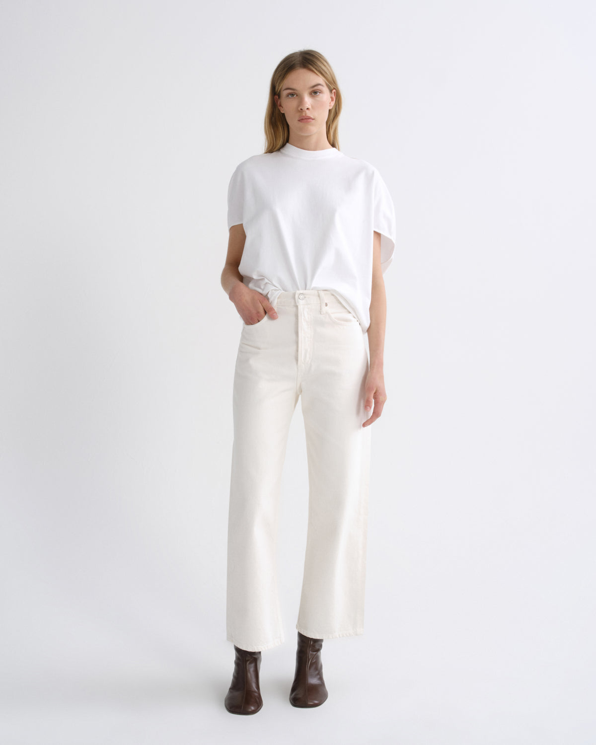 A model stands against a light gray studio background wearing a white short-sleeve t-shirt tucked into off-white wide-leg jeans, paired with dark brown ankle boots.