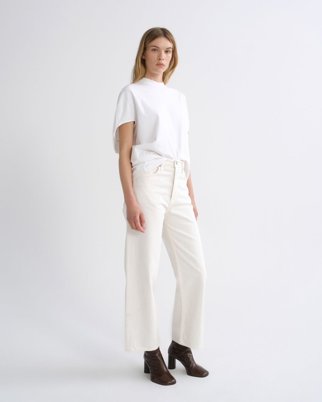 A model stands in a studio wearing a white short-sleeve top, off-white wide-leg cropped pants, and dark brown heeled ankle boots against a light gray background.