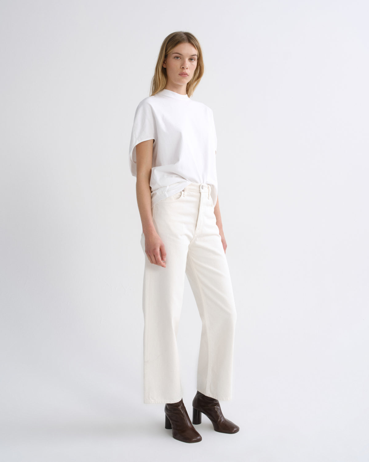 A model stands in a studio wearing a white short-sleeve top, off-white wide-leg cropped pants, and dark brown heeled ankle boots against a light gray background.