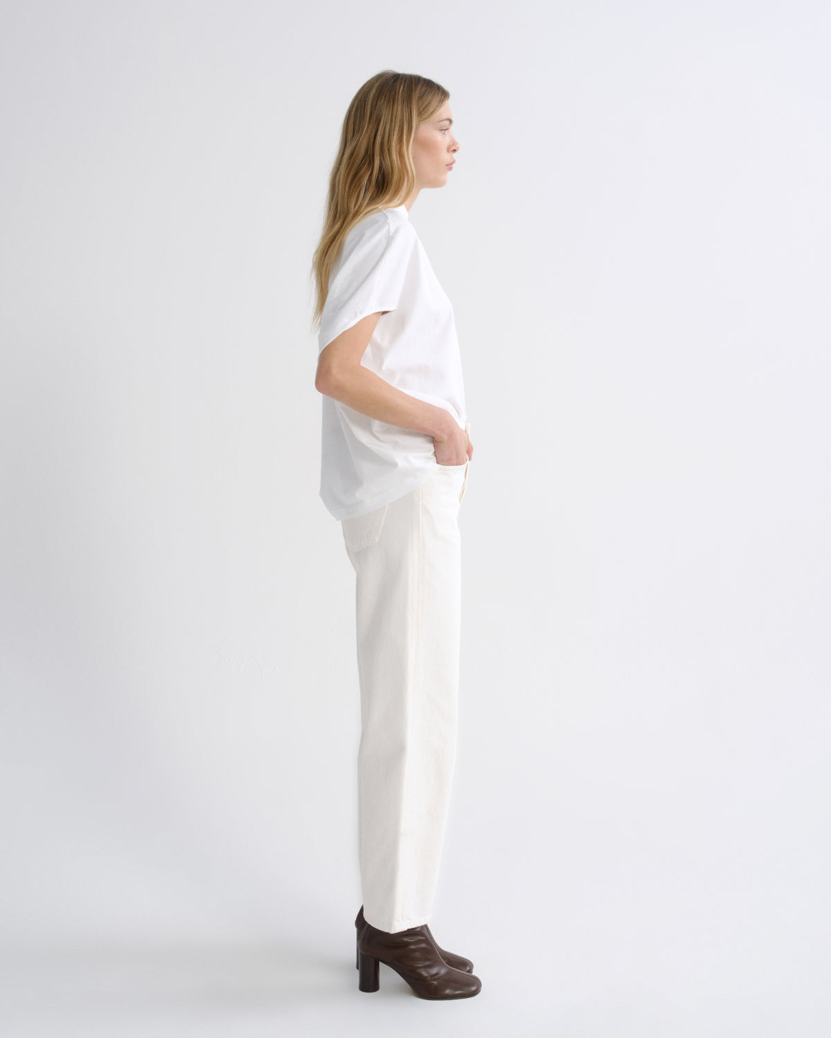 A woman shown in profile wearing a loose white short-sleeve top, white trousers, and dark brown heeled ankle boots against a plain light gray studio background.