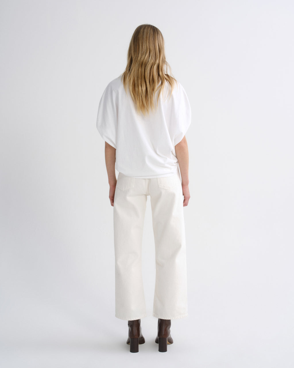 A woman viewed from behind wearing a loose white short-sleeve top and off-white wide-leg cropped pants with dark brown boots, set against a plain light gray studio background.