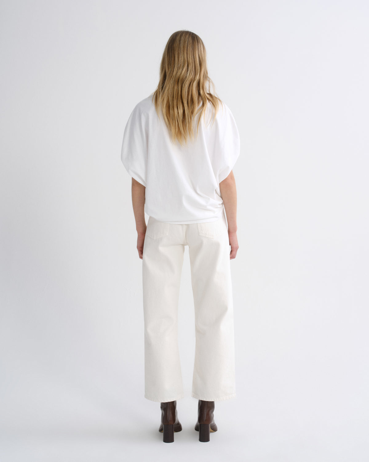 A woman viewed from behind wearing a loose white short-sleeve top and off-white wide-leg cropped pants with dark brown boots, set against a plain light gray studio background.