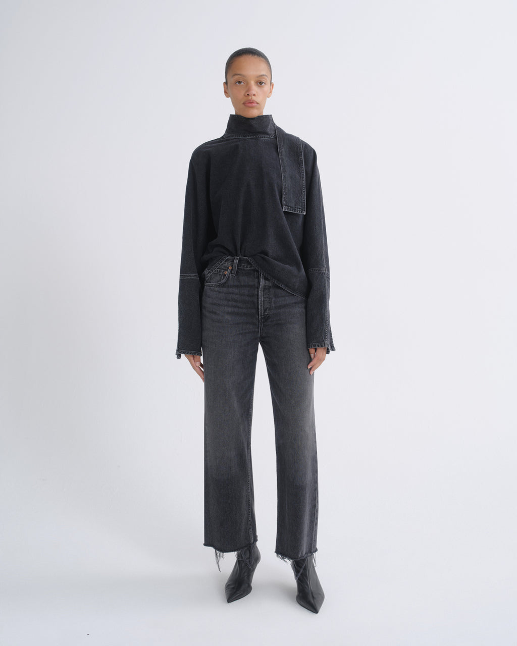 A model stands against a light gray studio background wearing a dark charcoal high-neck long-sleeve top, matching dark gray wide-leg jeans, and black pointed boots.