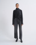 A model stands against a light gray studio background wearing a dark charcoal high-neck long-sleeve top, matching dark gray wide-leg jeans, and black pointed boots.