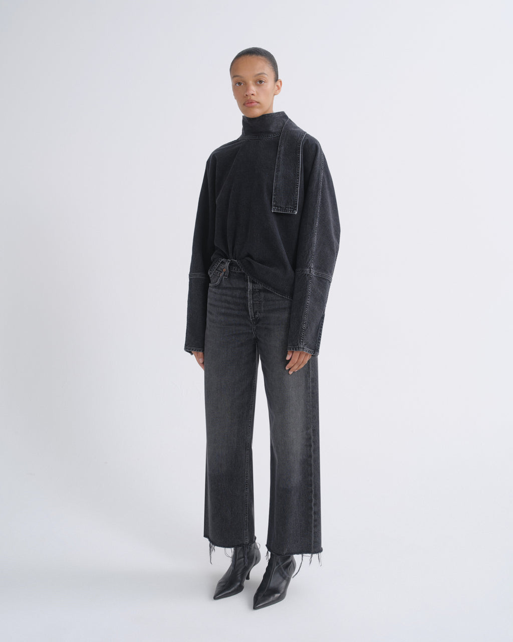 A model stands against a light gray studio background wearing a black long-sleeve mock-neck top with a draped pocket detail, dark gray wide-leg jeans with raw frayed hems, and black pointed ankle boots.