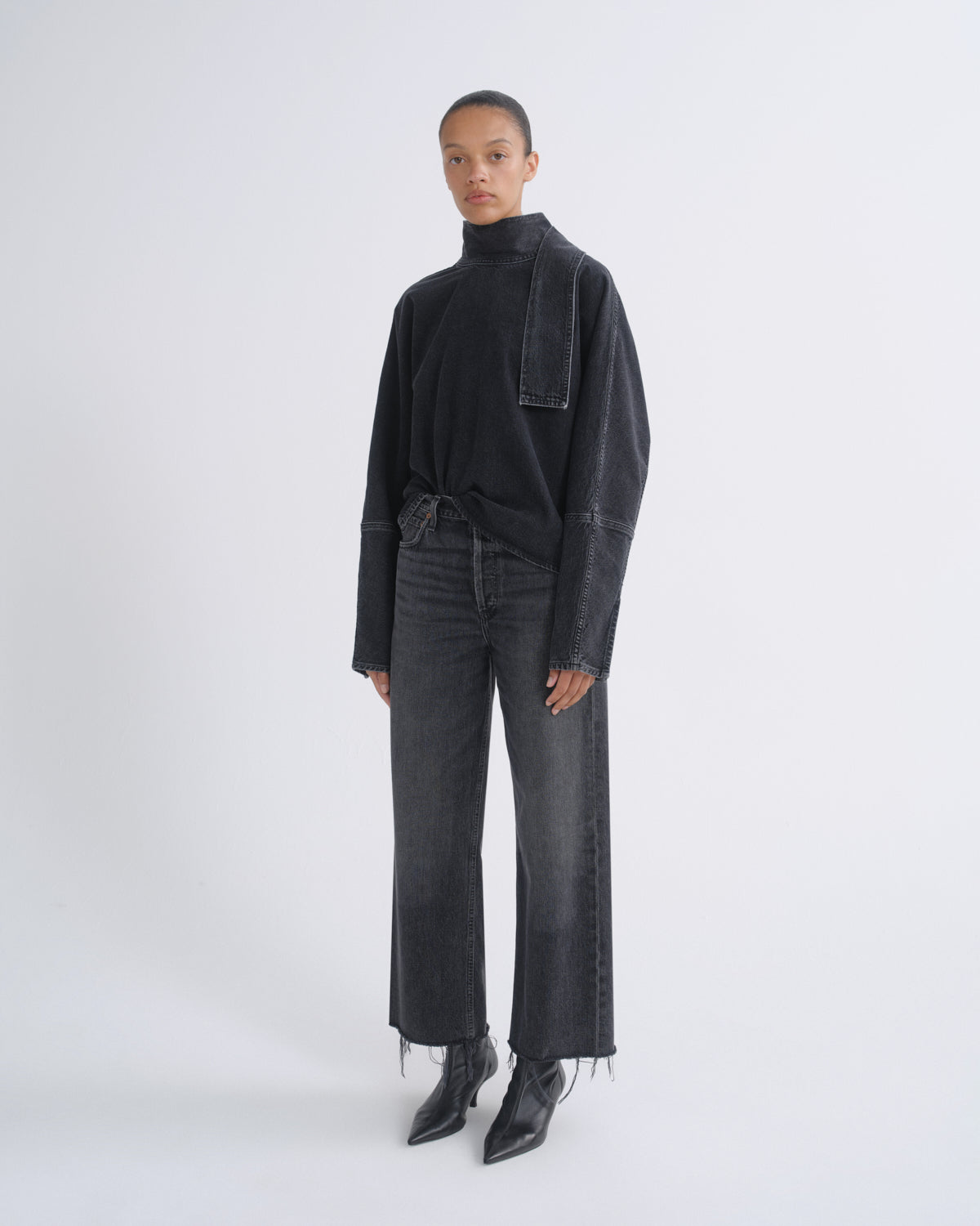 A model stands against a light gray studio background wearing a black long-sleeve mock-neck top with a draped pocket detail, dark gray wide-leg jeans with raw frayed hems, and black pointed ankle boots.