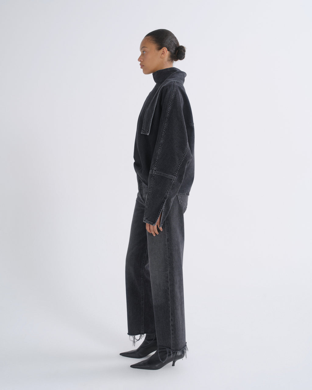 A model shown in profile wearing a dark washed denim jacket, matching wide-leg jeans, and black pointed-toe heeled boots against a plain light gray studio background.