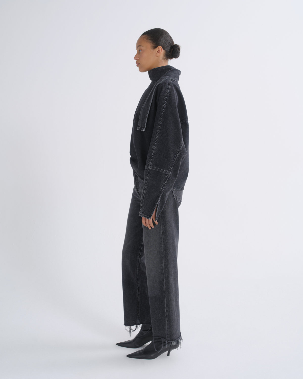 A model shown in profile wearing a dark washed denim jacket, matching wide-leg jeans, and black pointed-toe heeled boots against a plain light gray studio background.
