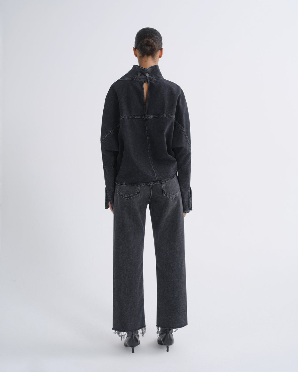 A model viewed from the back wearing a black long-sleeve high-neck top with a small open back detail and wide-leg dark gray jeans, standing against a light gray studio background.