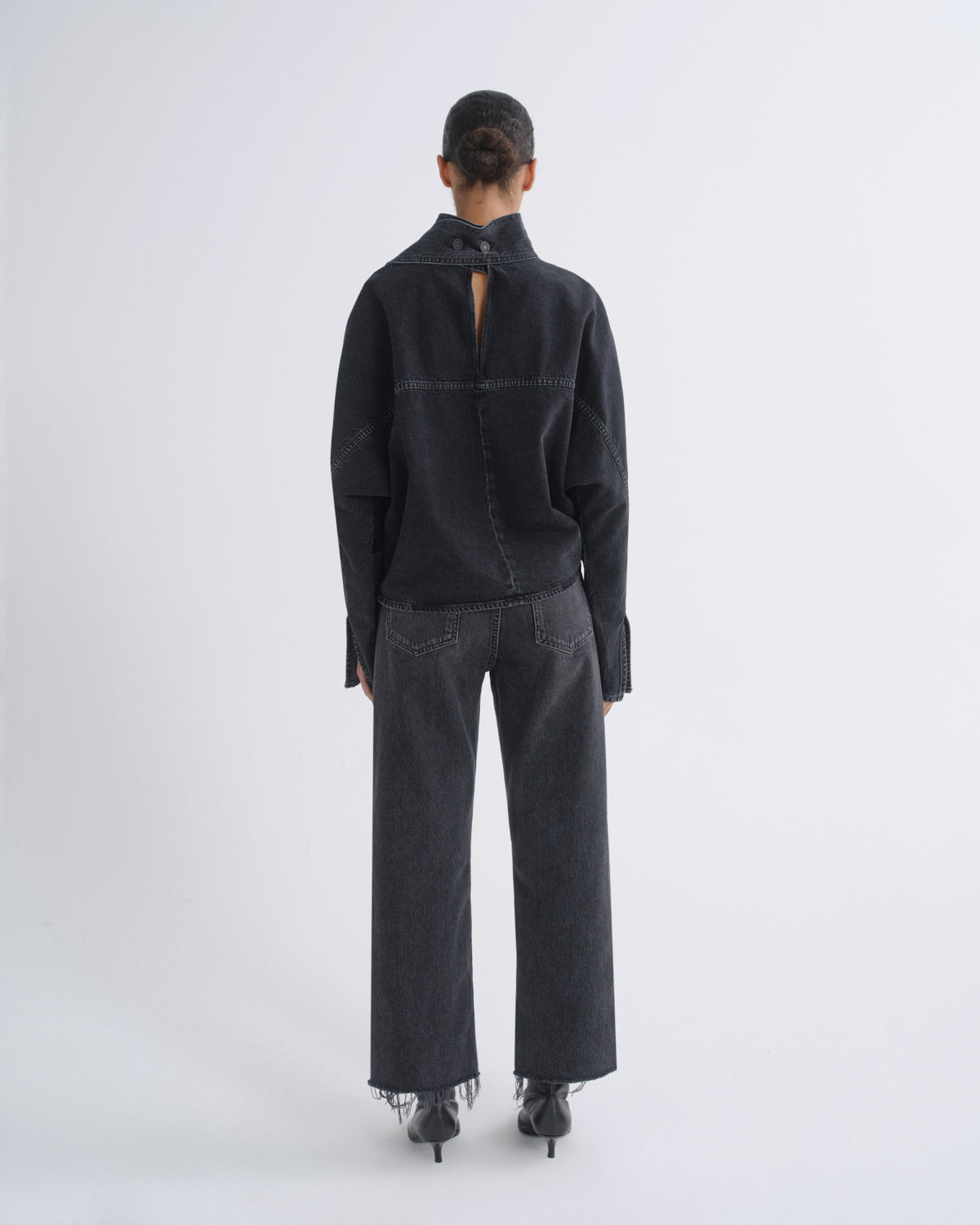 A model viewed from the back wearing a black long-sleeve high-neck top with a small open back detail and wide-leg dark gray jeans, standing against a light gray studio background.