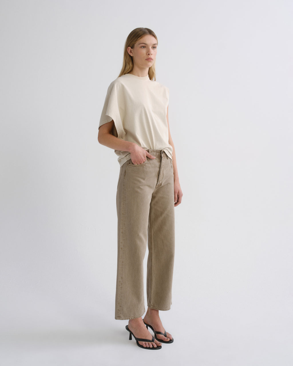 A woman stands in a studio wearing a cream short-sleeve top and beige wide-leg cropped pants with black heeled sandals against a light gray background.