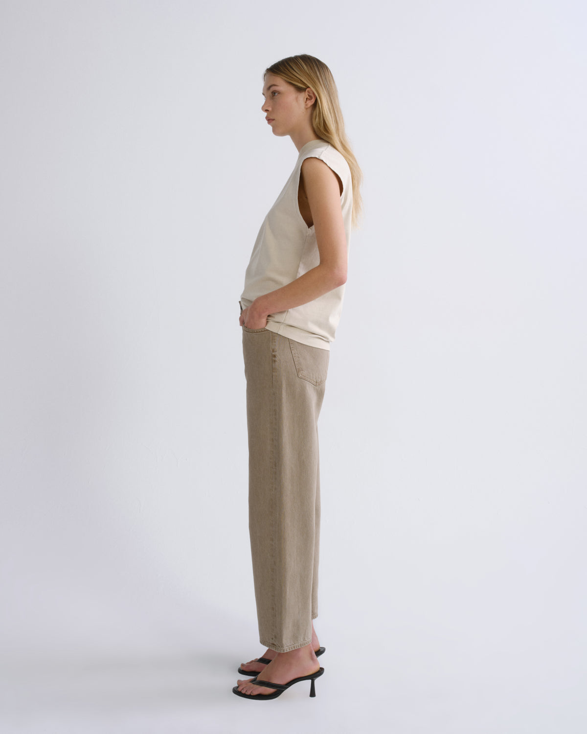 A woman stands in profile wearing a sleeveless cream top, beige wide-leg trousers, and black heeled sandals against a plain light gray studio background.