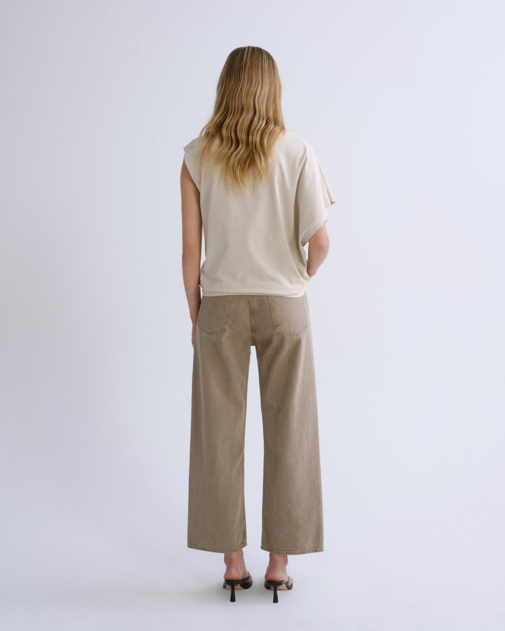 A woman viewed from behind wearing a cream-colored sleeveless top and beige wide-leg trousers, paired with black high-heeled sandals, against a light gray studio background.