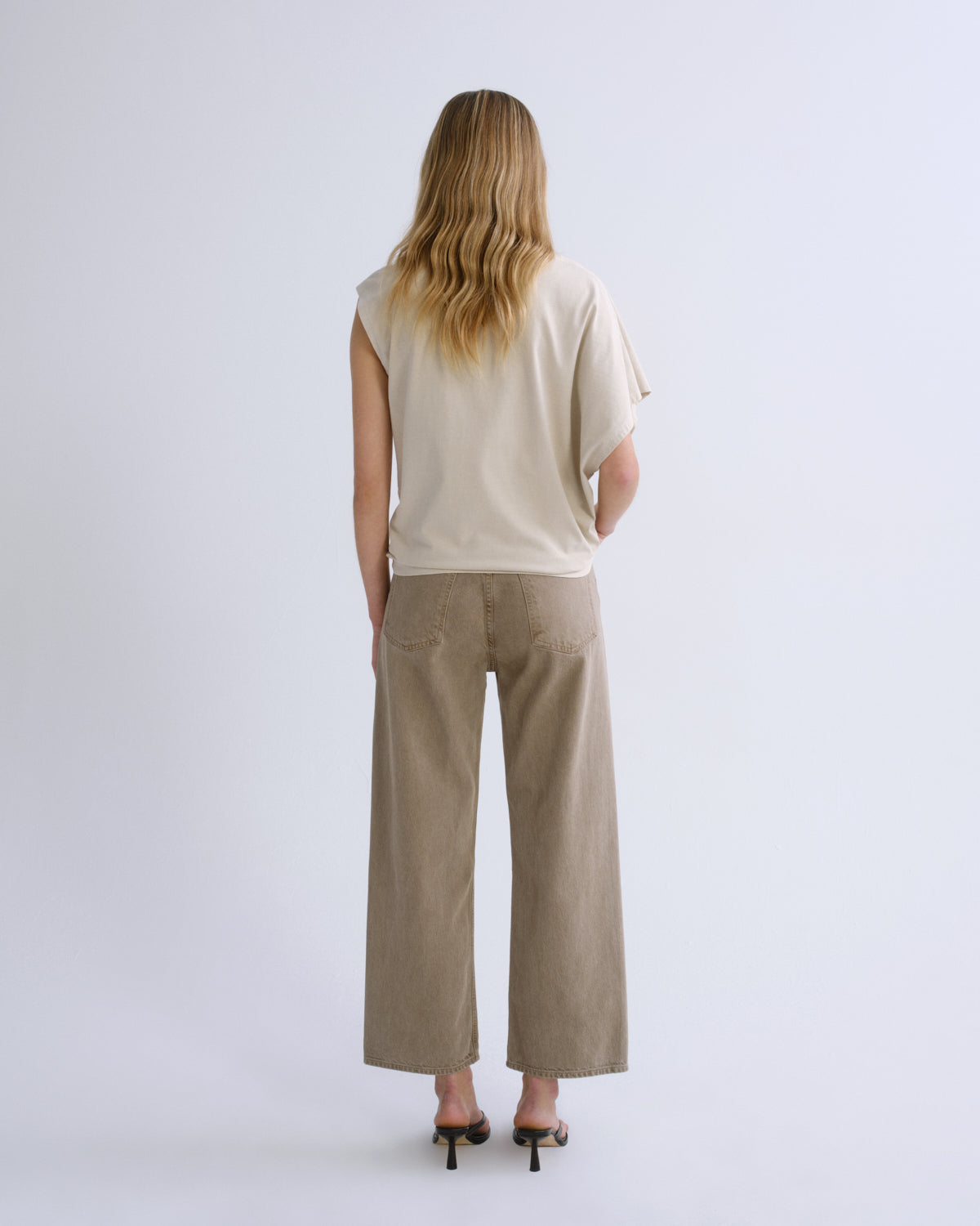A woman viewed from behind wearing a cream-colored sleeveless top and beige wide-leg trousers, paired with black high-heeled sandals, against a light gray studio background.