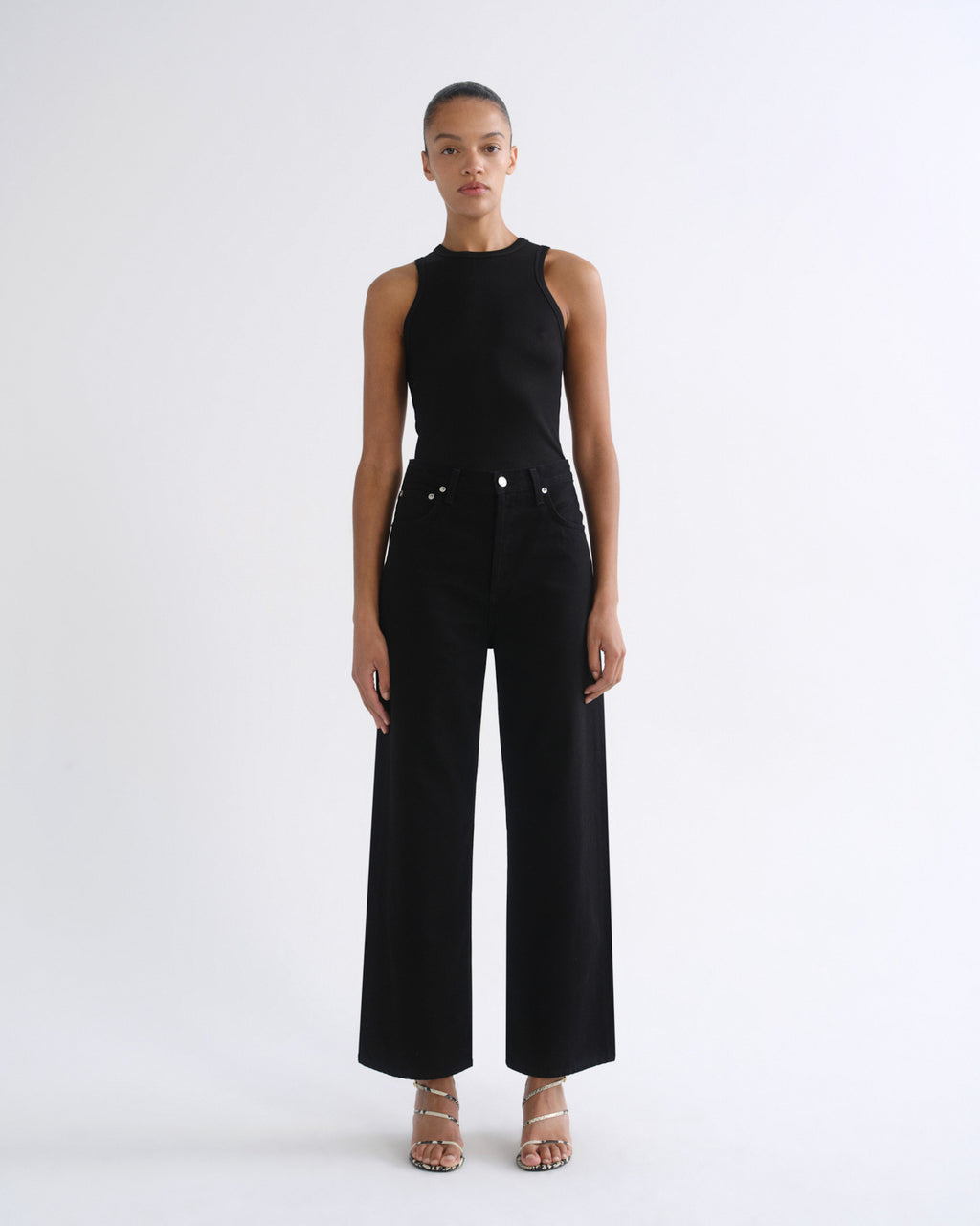 A model stands against a plain white studio background wearing a black sleeveless high-neck top and matching black wide-leg pants with silver-tone hardware, paired with clear-strap heeled sandals.