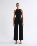 A model stands against a plain white studio background wearing a black sleeveless high-neck top and matching black wide-leg pants with silver-tone hardware, paired with clear-strap heeled sandals.