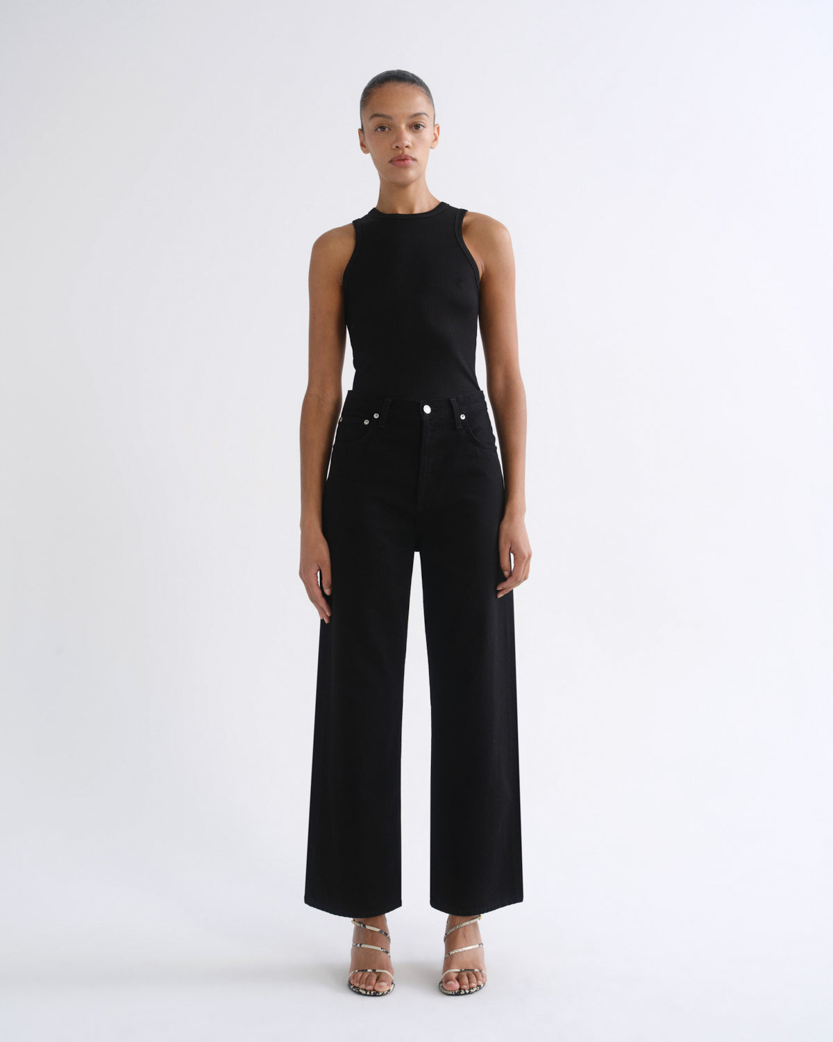 A model stands against a plain white studio background wearing a black sleeveless high-neck top and matching black wide-leg pants with silver-tone hardware, paired with clear-strap heeled sandals.