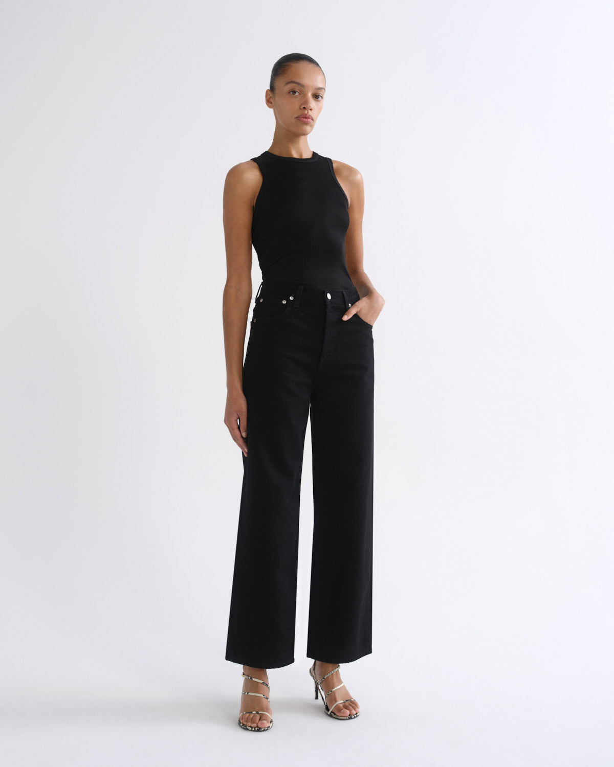 A model stands against a plain white studio background wearing a black sleeveless top and black wide-leg cropped pants, paired with metallic heeled sandals.