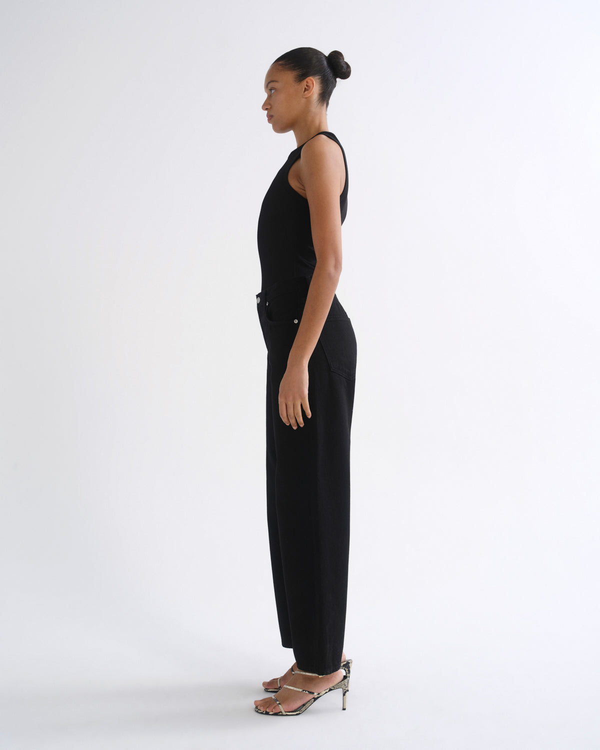 A woman in a black sleeveless jumpsuit and metallic heeled sandals, shown in profile against a plain white studio background.