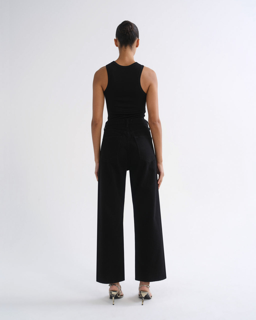 A model viewed from the back wearing a black sleeveless top and black wide-leg trousers, paired with metallic high-heeled sandals, against a plain white studio background.
