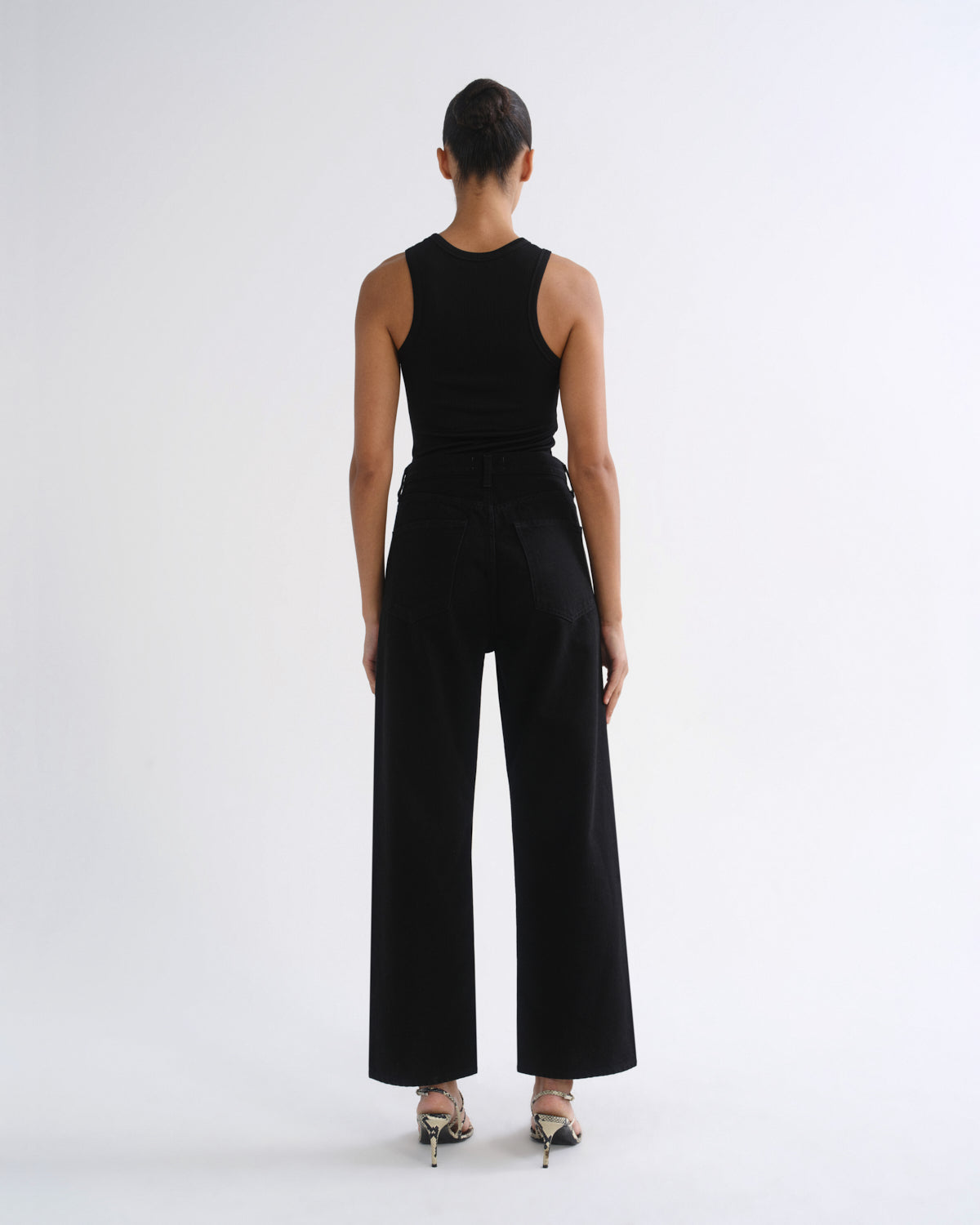 A model viewed from the back wearing a black sleeveless top and black wide-leg trousers, paired with metallic high-heeled sandals, against a plain white studio background.