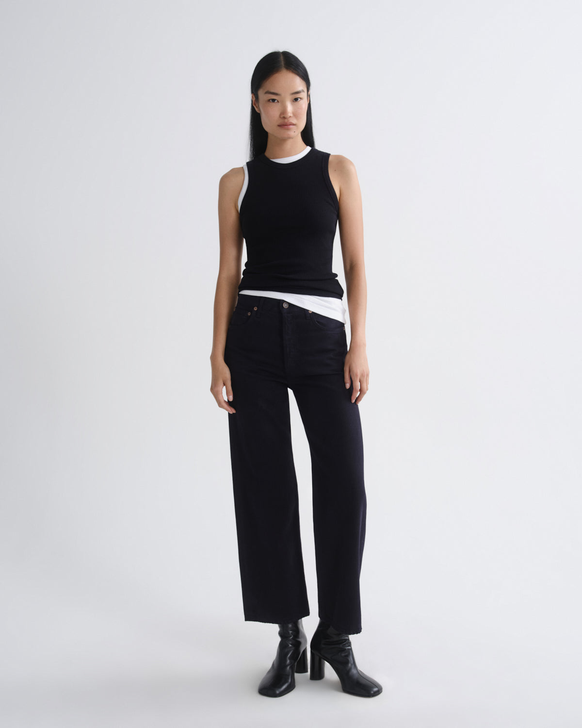 A model stands against a light gray studio background wearing a black sleeveless top with a white inner collar and hem, black wide-leg cropped pants, and black heeled ankle boots.