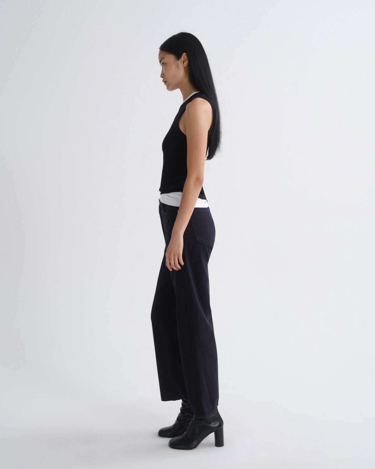 A woman stands in profile wearing a black sleeveless top, dark wide-leg pants, and black heeled boots against a plain light gray studio background.