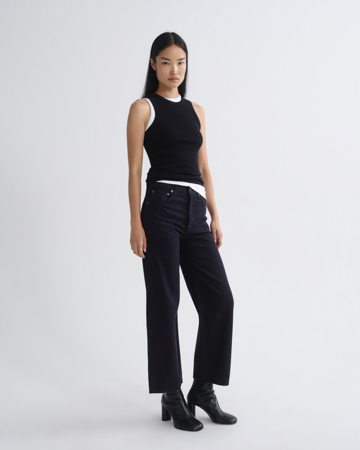 A model stands against a light gray studio background wearing a black sleeveless top with a white collar, dark wide-leg pants, and black ankle boots.