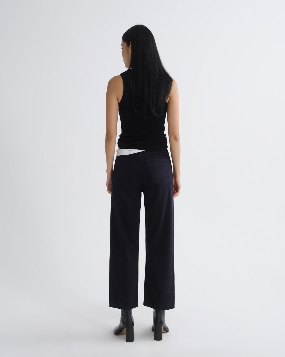A model viewed from the back wearing a black sleeveless top, black wide-leg cropped trousers, and black ankle boots against a plain light gray studio background.