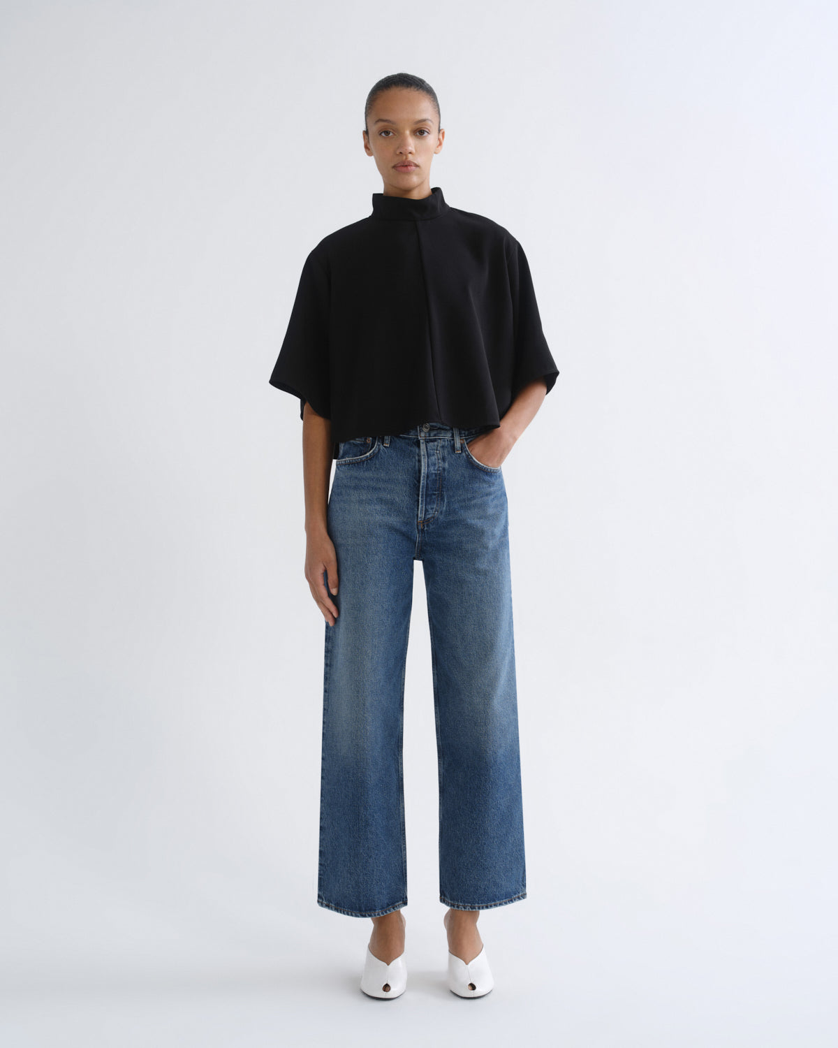 A model stands against a light gray studio background wearing a black short-sleeve mock-neck top, wide-leg blue jeans, and white pointed shoes.