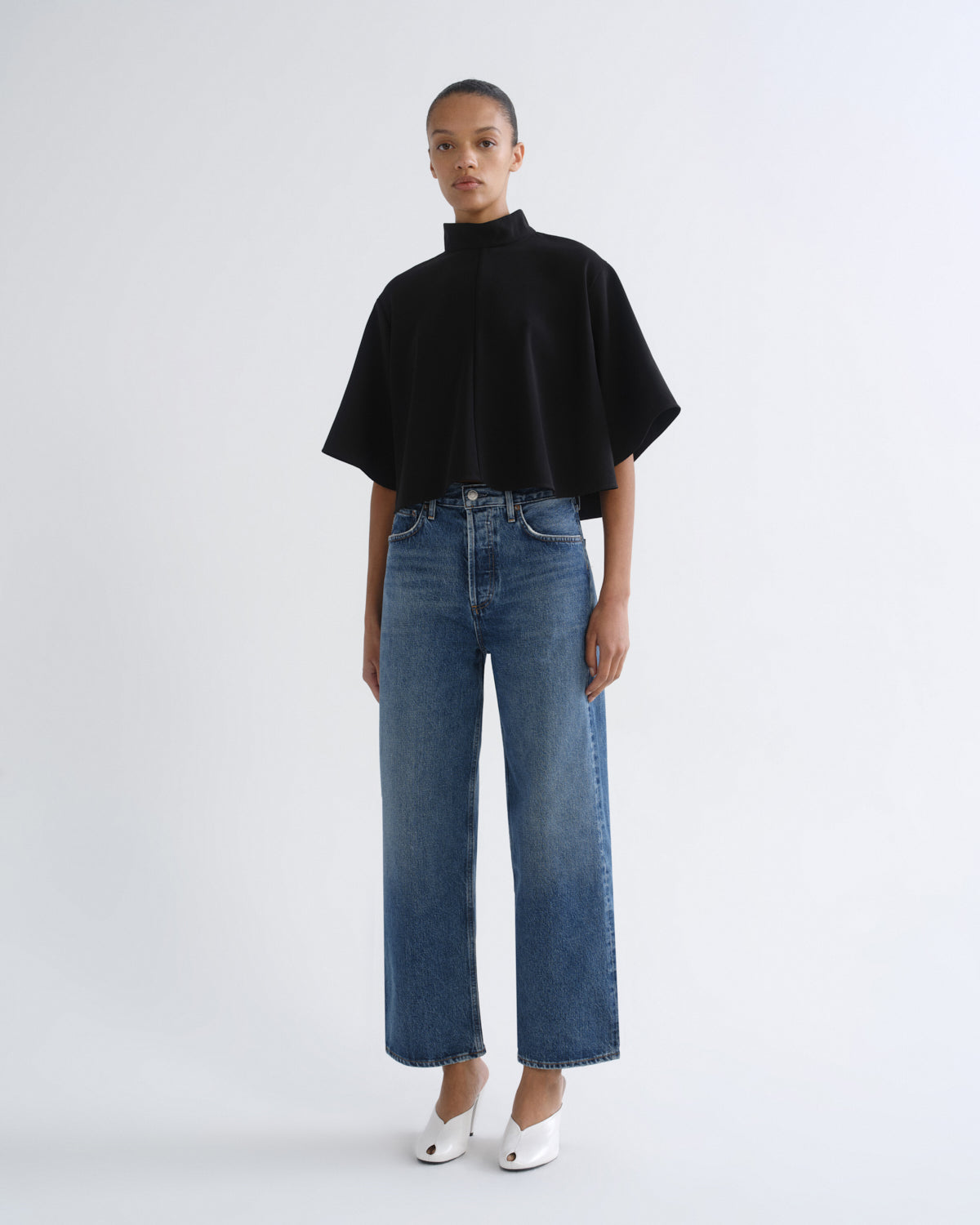A model stands against a light gray studio background wearing a black short-sleeved mock-neck top, blue wide-leg cropped jeans, and white pointed shoes.