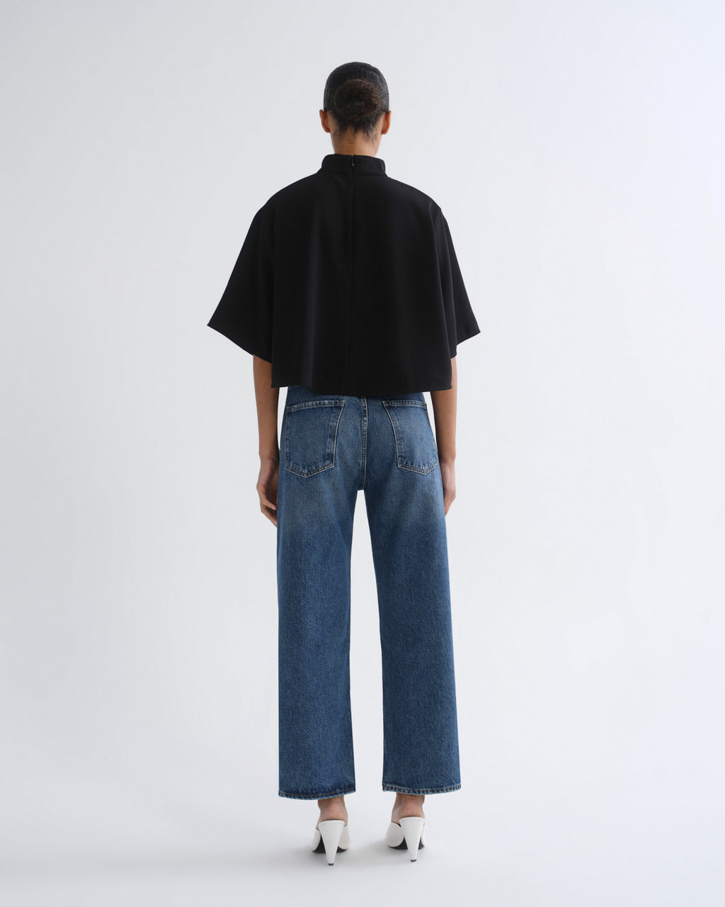 A model shown from the back wearing a black short-sleeve cropped top, wide-leg blue jeans, and white pointed-toe heels against a plain light gray studio background.