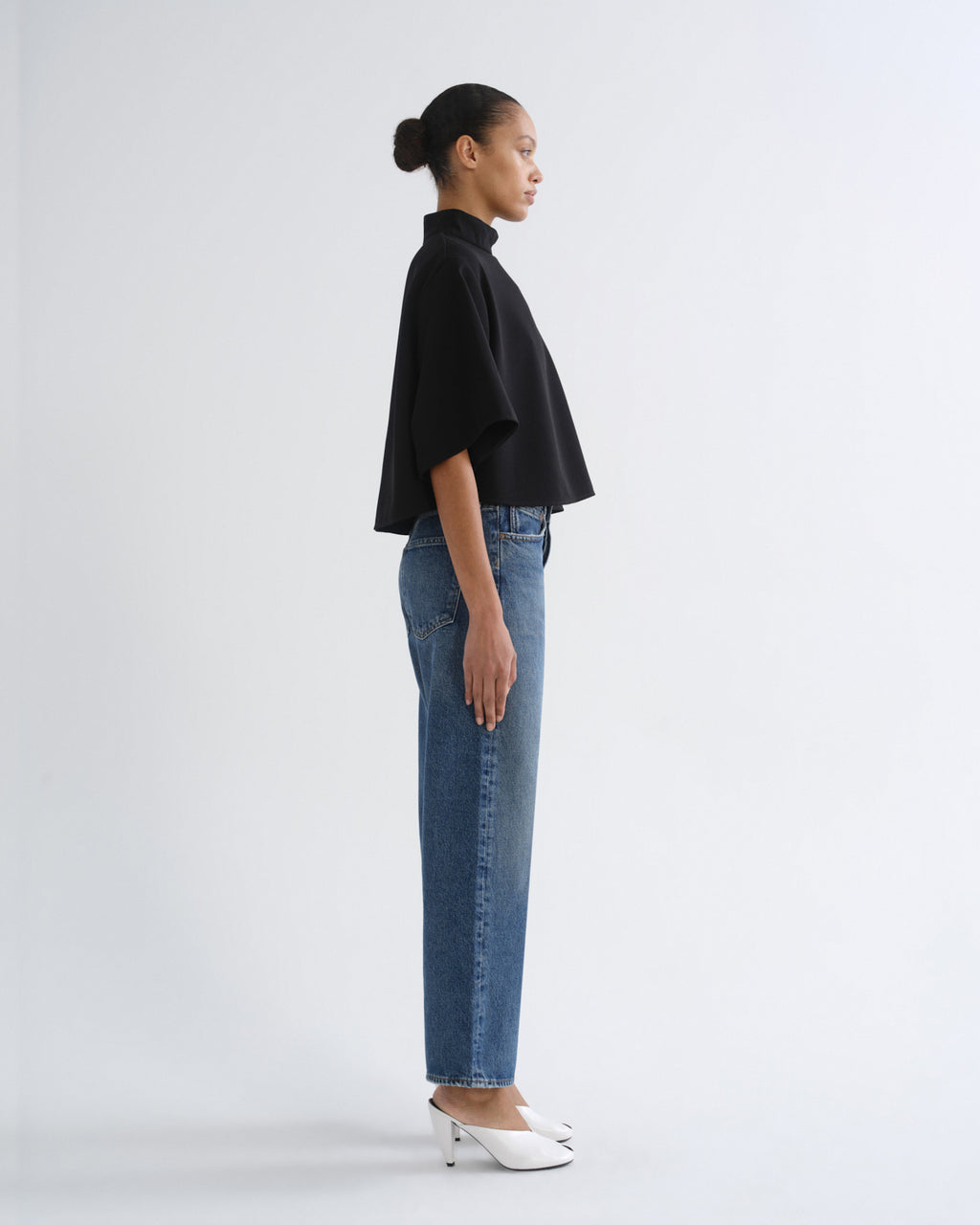 A model shown in profile wearing a black short-sleeve mock-neck top, blue straight-leg jeans, and white pointed-toe heels against a plain light gray studio background.