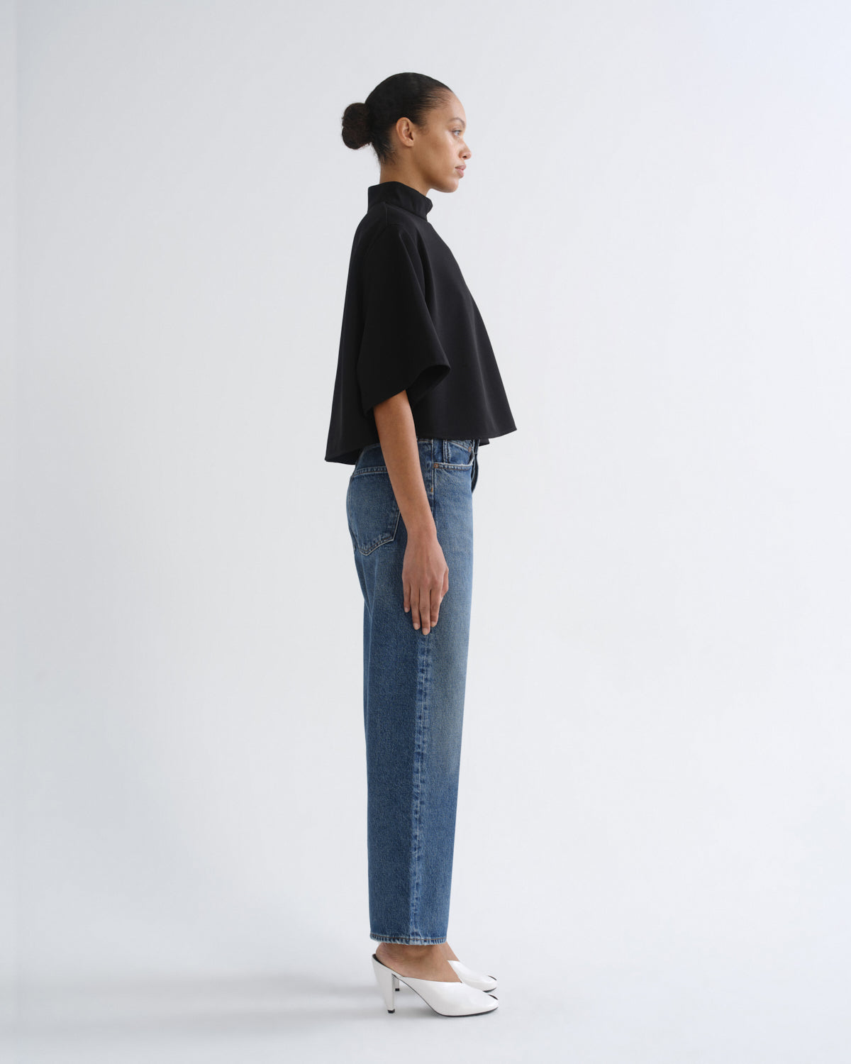 A model shown in profile wearing a black short-sleeve mock-neck top, blue straight-leg jeans, and white pointed-toe heels against a plain light gray studio background.